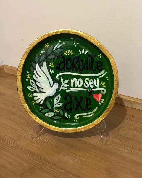 Hand-painted decorative plate on a stand, deep green with a gold rim, showing a white dove with olive branches and the hand-lettered words "Acredite no seu axé".
