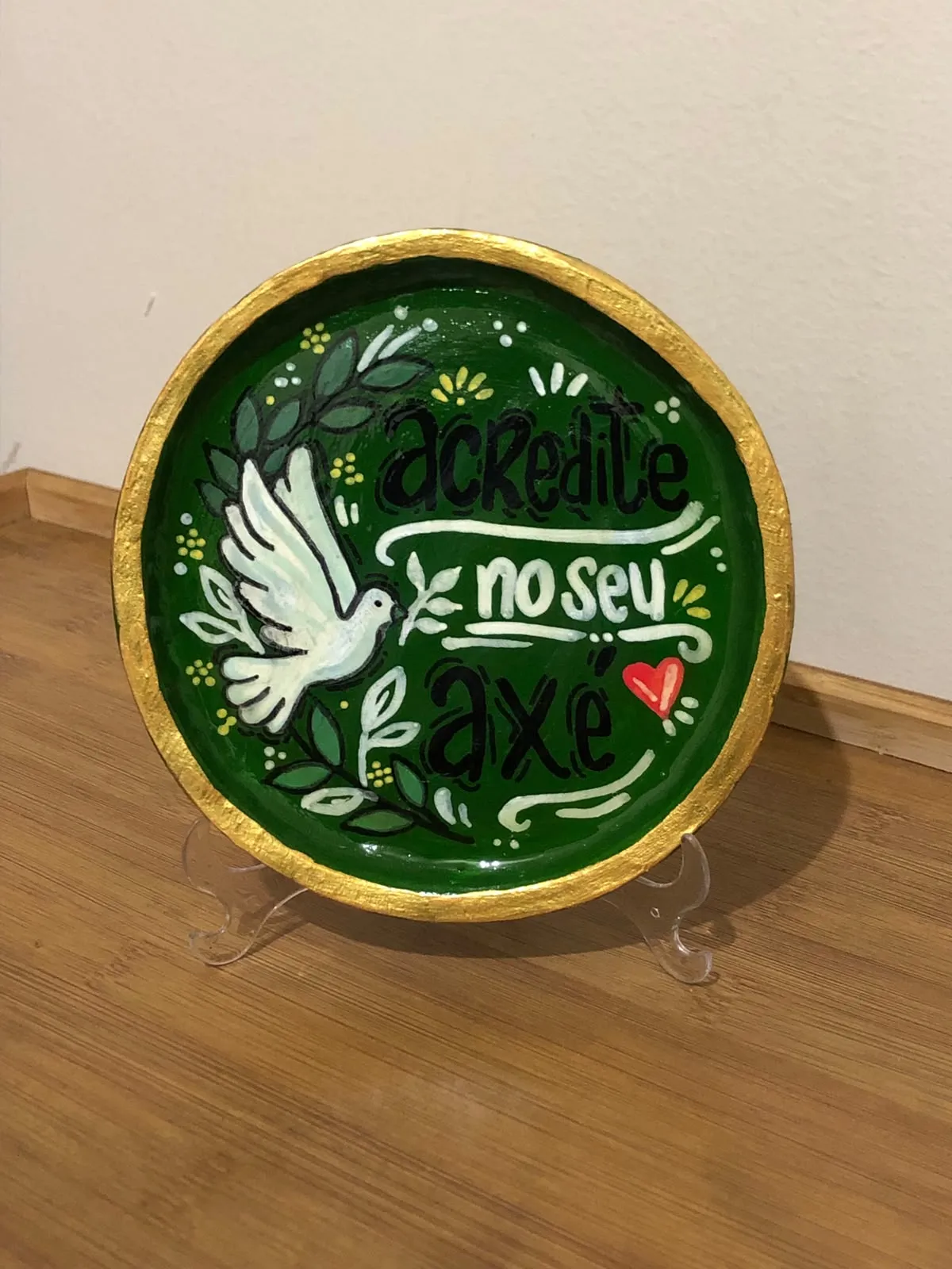 Hand-painted decorative plate on a stand, deep green with a gold rim, showing a white dove with olive branches and the hand-lettered words "Acredite no seu axé".