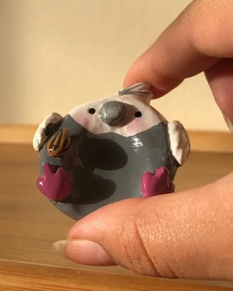Tiny hand-sculpted grey cockatiel figurine with pink cheeks holding a sunflower seed, held between two fingers.