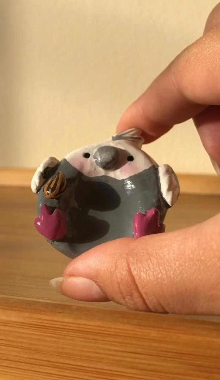 Tiny hand-sculpted grey cockatiel figurine with pink cheeks holding a sunflower seed, held between two fingers.