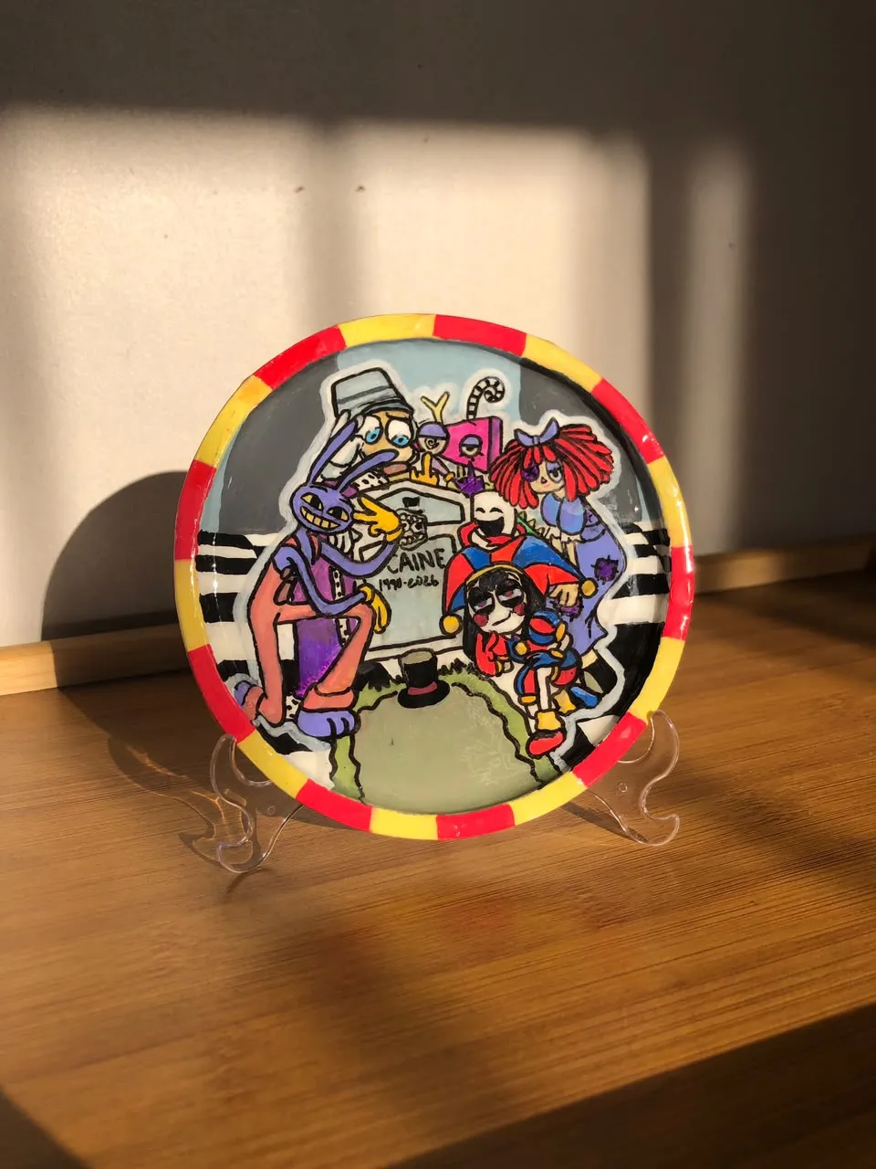 Hand-painted decorative plate on a stand with a red and yellow rim showing The Amazing Digital Circus characters gathered around a tombstone.