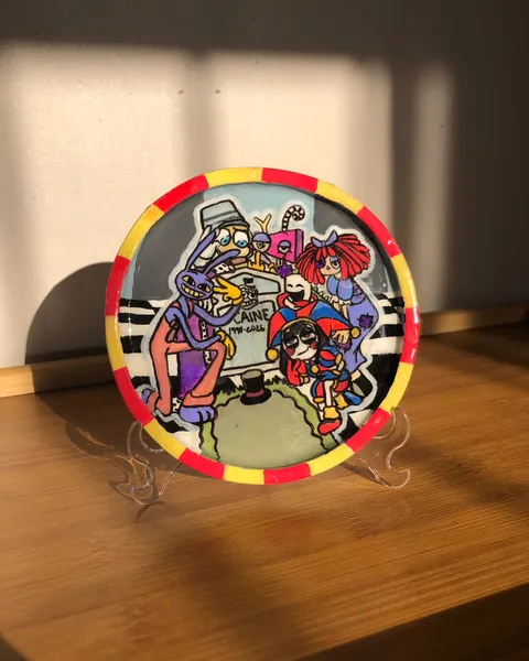 Hand-painted decorative plate on a stand with a red and yellow rim showing The Amazing Digital Circus characters gathered around a tombstone.