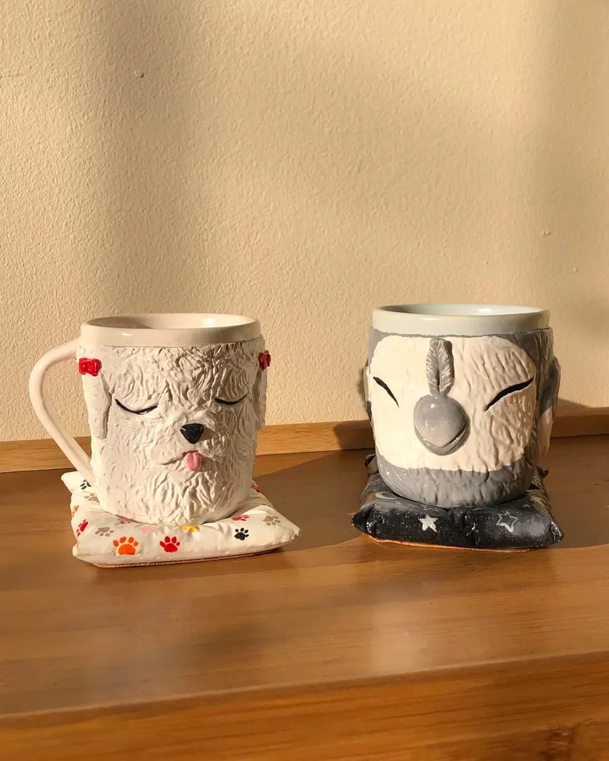 Two sculpted mugs shaped like a sleeping curly white dog with red bows and a sleeping grey cockatiel, each resting on a small pillow coaster on a wooden table.