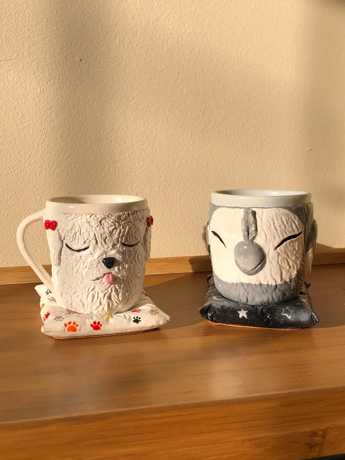 Two sculpted mugs shaped like a sleeping curly white dog with red bows and a sleeping grey cockatiel, each resting on a small pillow coaster on a wooden table.