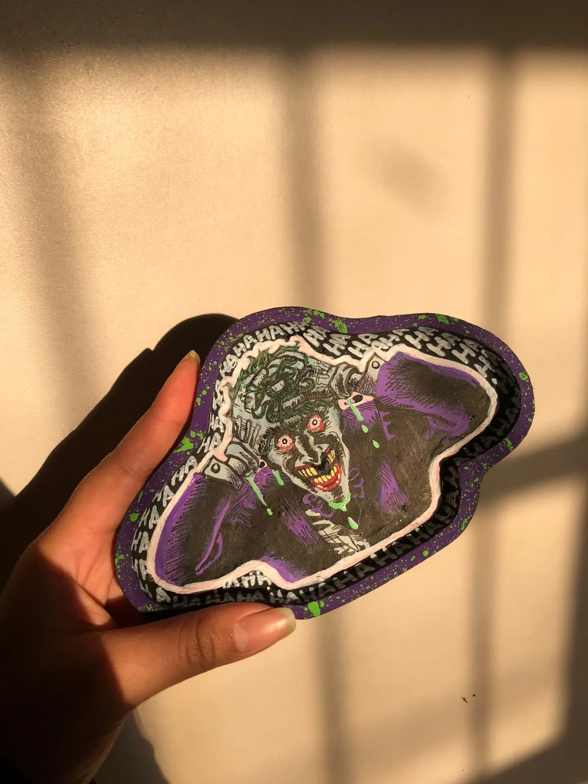 Hand holding a free-form purple trinket tray painted with a laughing Joker portrait and a HAHAHA border, lit by afternoon sun.