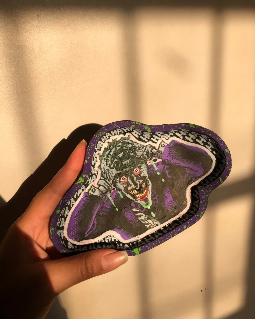 Hand holding a free-form purple trinket tray painted with a laughing Joker portrait and a HAHAHA border, lit by afternoon sun.