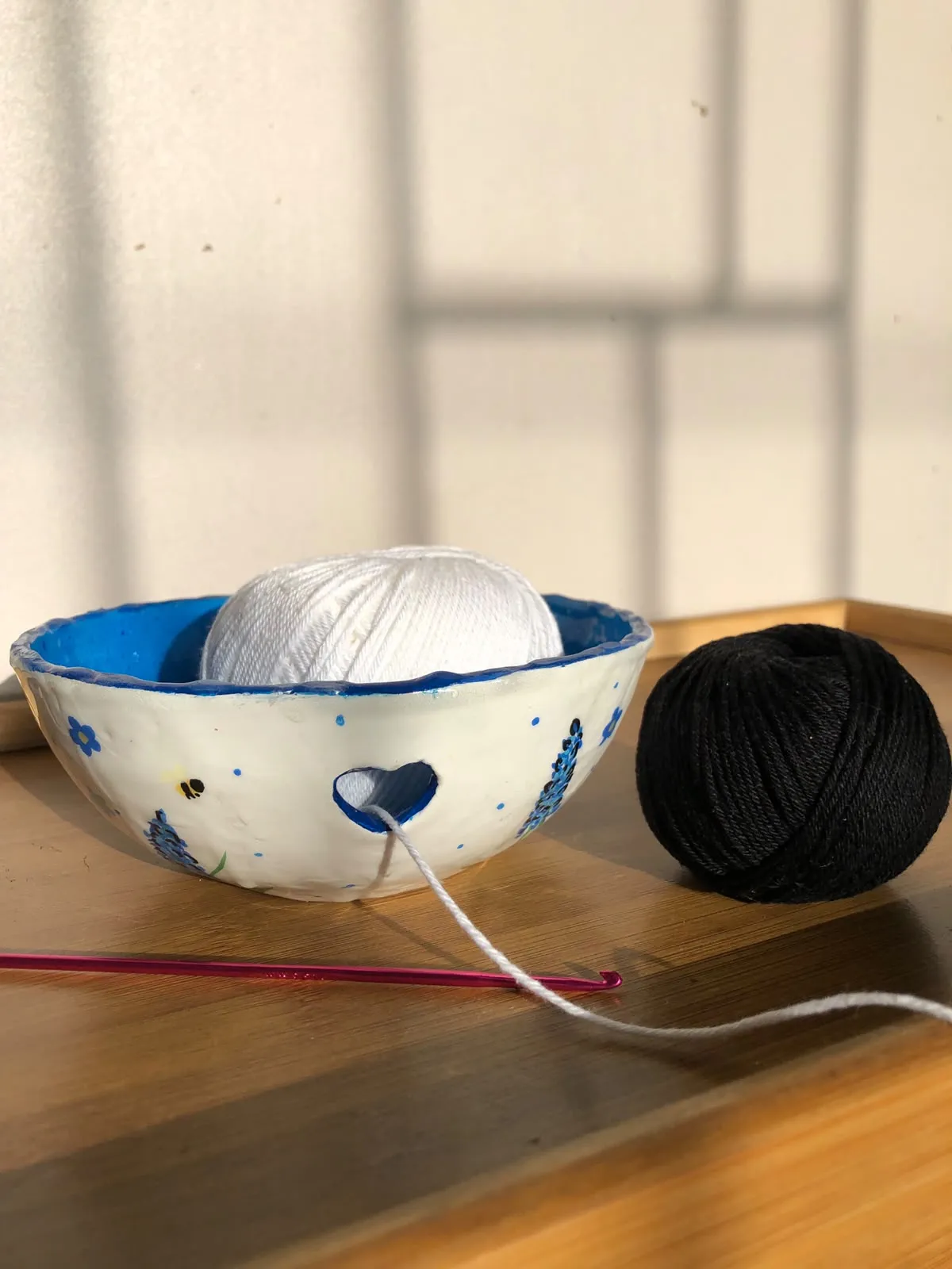 Hand-sculpted white yarn bowl with a heart-shaped cut-out, painted with lavender and bees, holding a white ball of yarn beside a black one and a pink crochet hook.