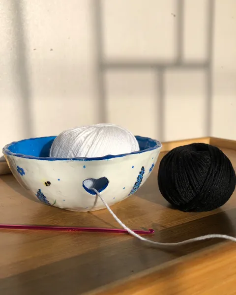 Hand-sculpted white yarn bowl with a heart-shaped cut-out, painted with lavender and bees, holding a white ball of yarn beside a black one and a pink crochet hook.