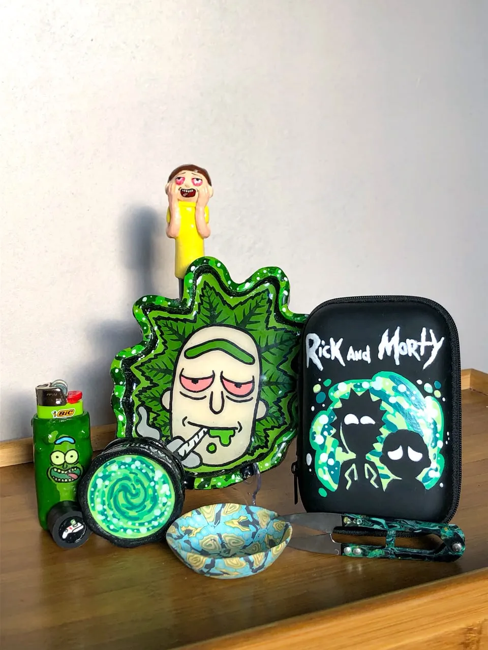 Hand-painted Rick and Morty themed set on a wooden table, a green Rick tray, portal grinder, Pickle Rick lighter, black painted case and a small Morty figure.