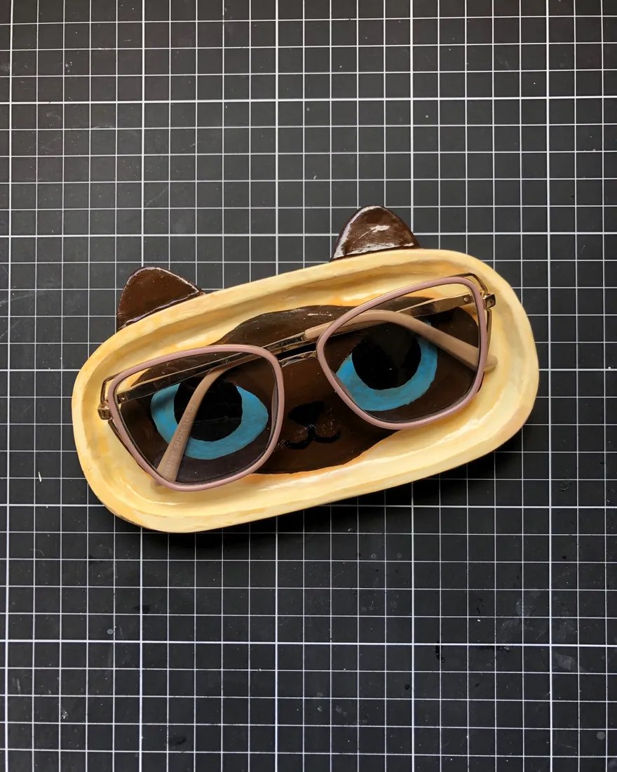Oval hand-sculpted trinket tray painted as a Siamese cat face with blue eyes and brown ears, holding a pair of glasses, on a black cutting mat.