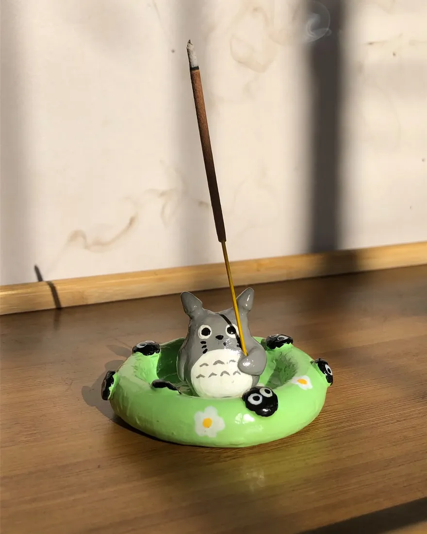Hand-sculpted incense holder shaped like Totoro sitting in a green pool float with soot sprites, holding a burning incense stick, on a wooden table in sunlight.
