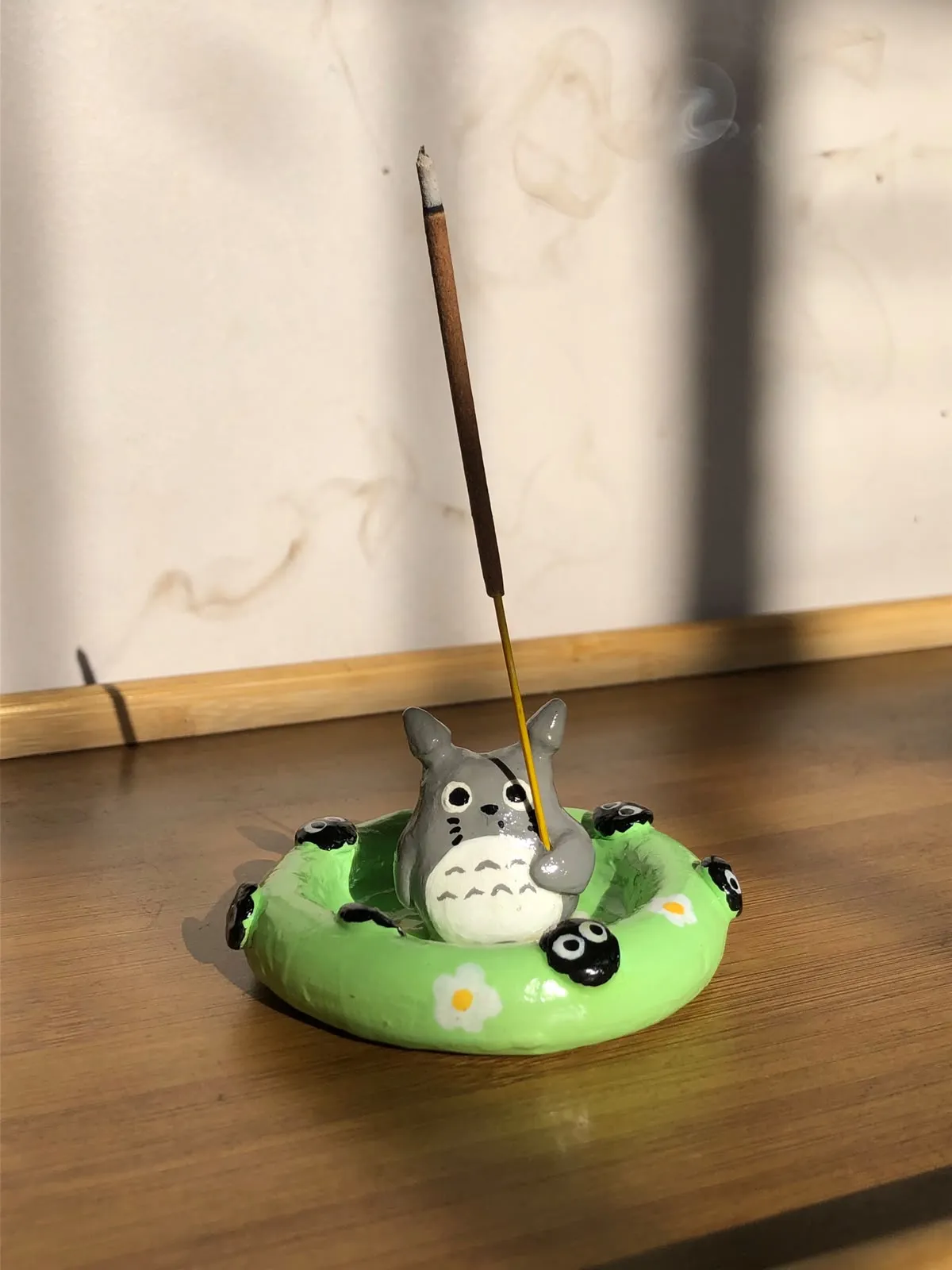 Hand-sculpted incense holder shaped like Totoro sitting in a green pool float with soot sprites, holding a burning incense stick, on a wooden table in sunlight.