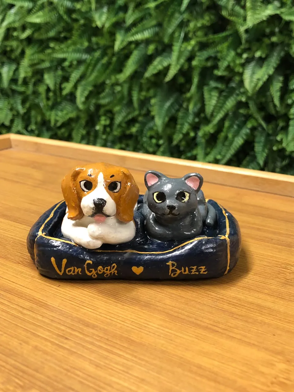 Miniature clay sculpture of a beagle and a grey cat lying together in a navy blue pet bed with the names Van Gogh and Buzz painted in gold.