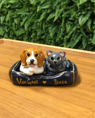 Miniature clay sculpture of a beagle and a grey cat lying together in a navy blue pet bed with the names Van Gogh and Buzz painted in gold.